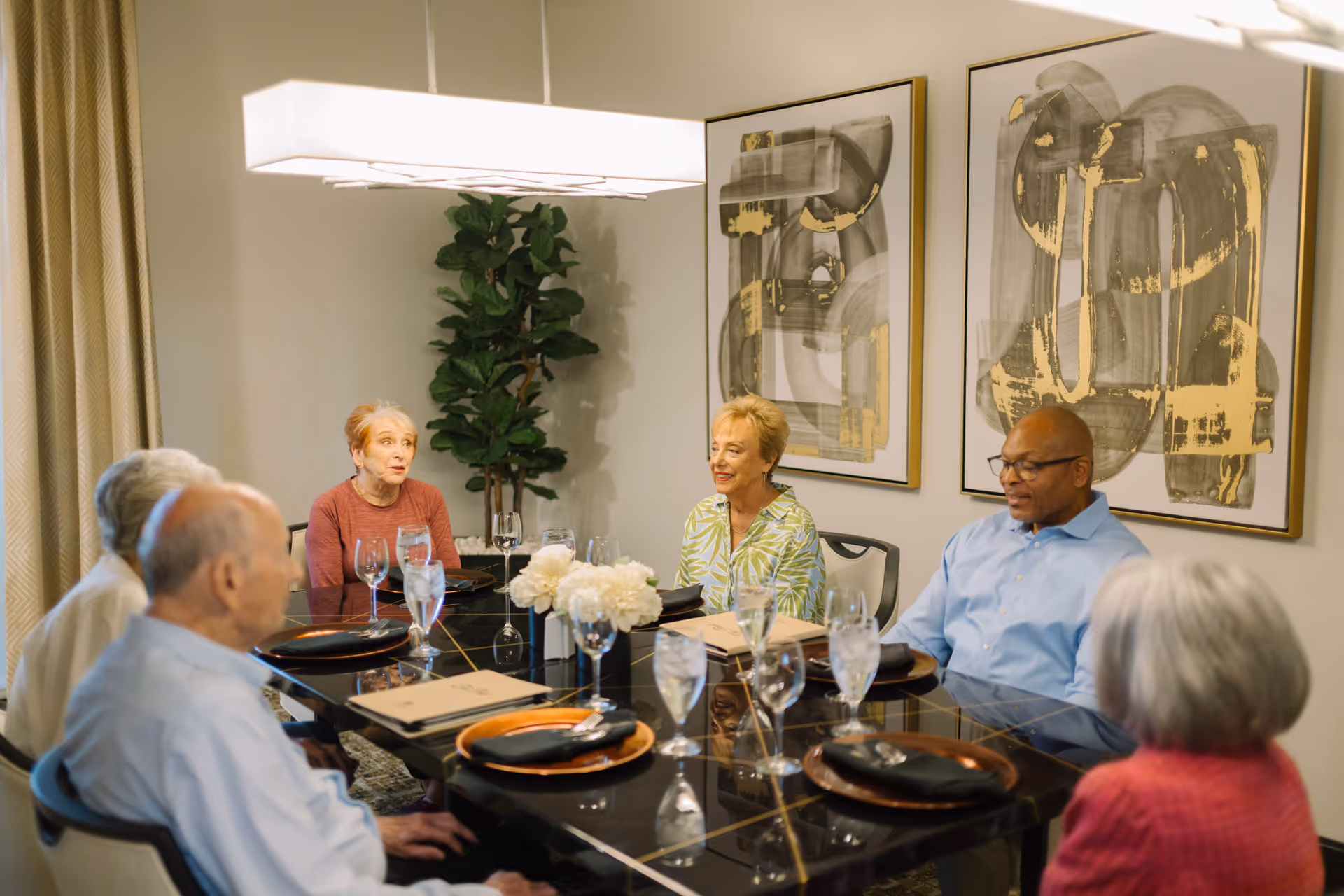 Several older adults seated around a formal dining table set with plates and glasses in a bright dining room.