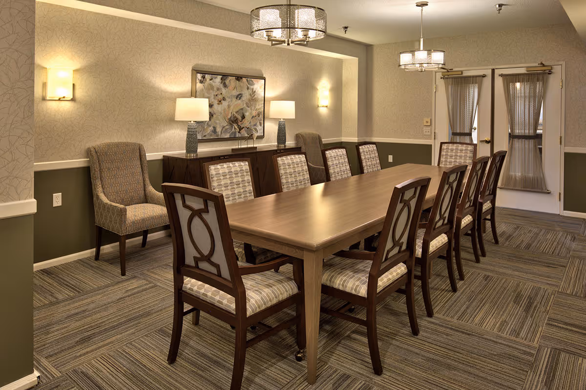 A well-lit dining room with a long wooden table surrounded by ten upholstered chairs. The room features patterned carpet, two modern chandeliers, wall sconces, a sideboard with two table lamps, and a large abstract painting on the wall. Double doors with sheer curtains are visible in the background.