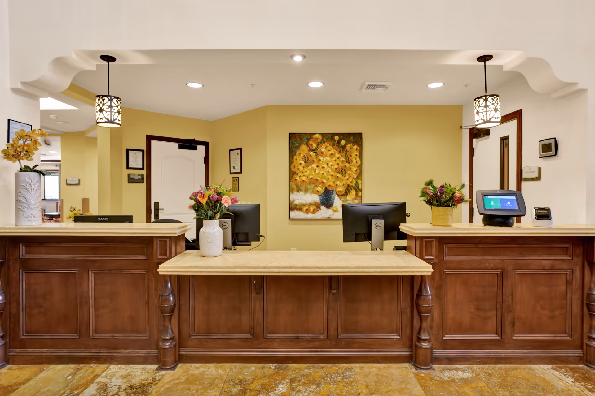 Reception desk area in a senior living facility with wooden counters, two computer monitors, decorative flower arrangements, and a painting of sunflowers on a yellow wall behind the desk.