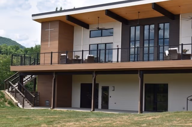 Modern two-story building facade with an upper deck and outdoor seating overlooking a grassy lawn.