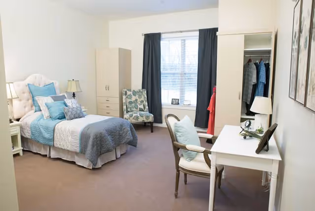 A neatly arranged bedroom with a single bed made with blue and gray bedding, a white tufted headboard, a bedside table with a lamp, a floral patterned armchair near a window with dark curtains, a white desk with a chair, and an open closet with hanging clothes.