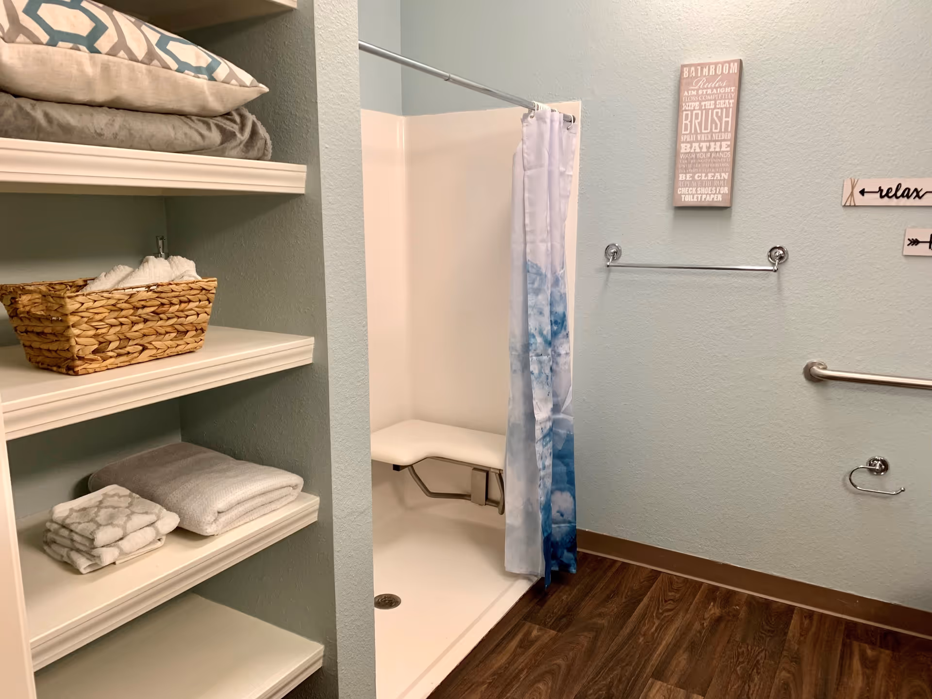 A bathroom with a walk-in shower featuring a fold-down seat and a blue and white shower curtain. There are shelves on the left side holding folded towels and a woven basket. The walls are light blue with decorative signs, and there are metal grab bars and a towel rack installed. The floor has a wood-like finish.