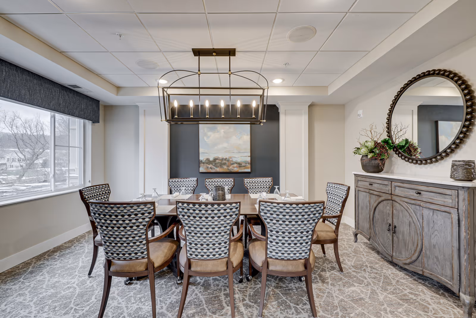 Well-appointed dining room with a rectangular table surrounded by patterned chairs, a large window, chandelier, and a sideboard with a round mirror.