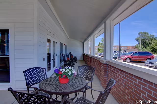 Covered outdoor patio area with a round metal table and four chairs, a red flower pot with colorful flowers on the table, several black rocking chairs lined up against the white wall with windows and doors, and a view of parked cars and trees in the background under a clear blue sky.