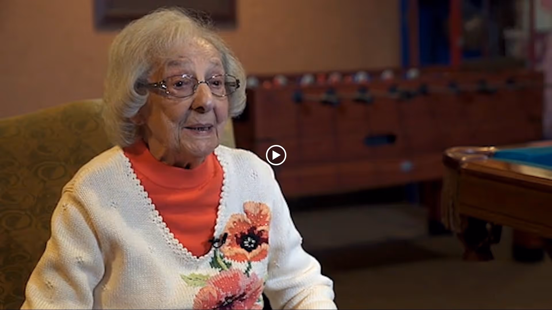 An elderly woman wearing glasses and a white sweater with a floral design is sitting in a chair in a room with a foosball table and a pool table in the background.