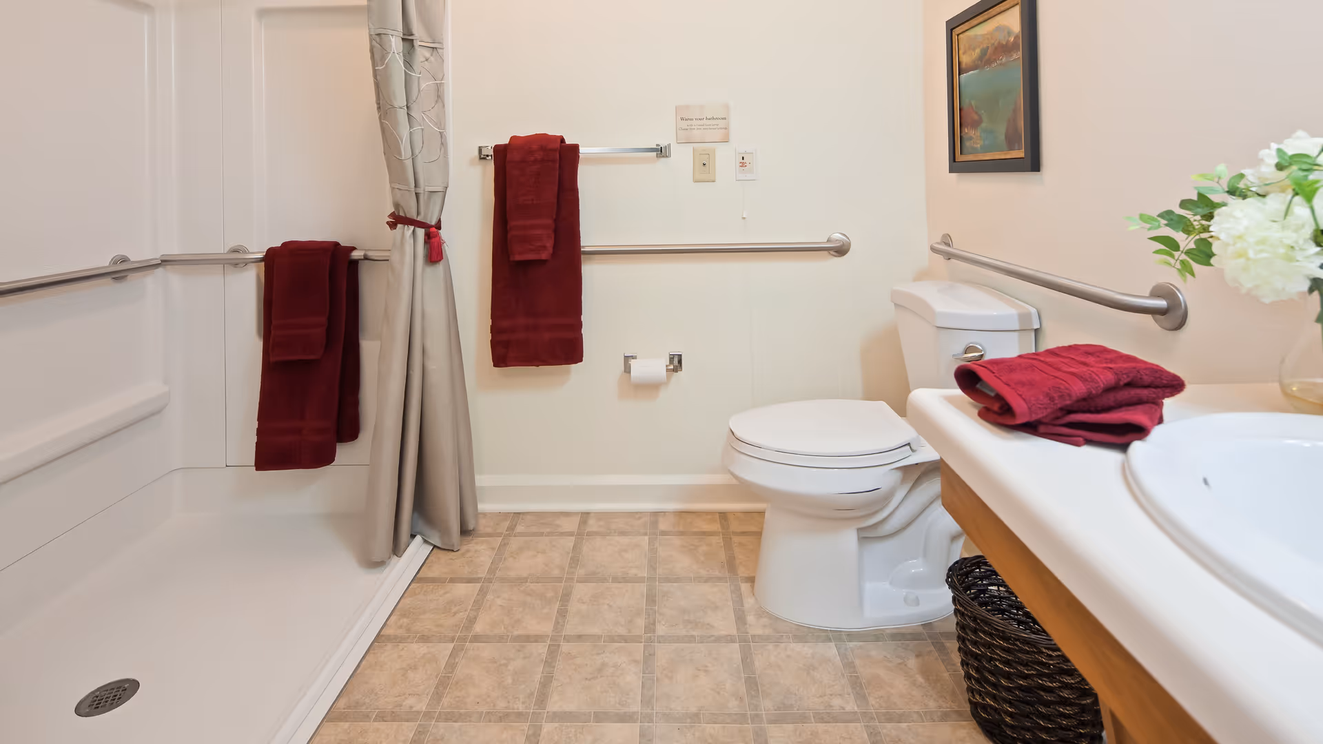 A clean and accessible bathroom with a walk-in shower on the left, equipped with grab bars and red towels hanging on the bars. There is a toilet in the center-right with a grab bar behind it, and a sink with a countertop on the right side, holding folded red towels and a vase with white flowers. The floor has a beige tile pattern, and a framed picture hangs on the wall above the toilet.