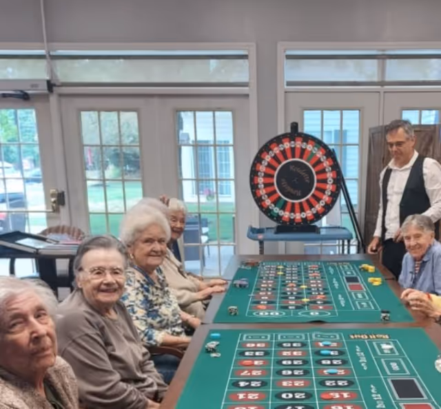 A group of elderly people sitting around a green casino-style gaming table inside a room with large windows and glass doors. A man in a white shirt and black vest stands nearby, and a large spinning wheel labeled 'Roulette' is visible in the background.