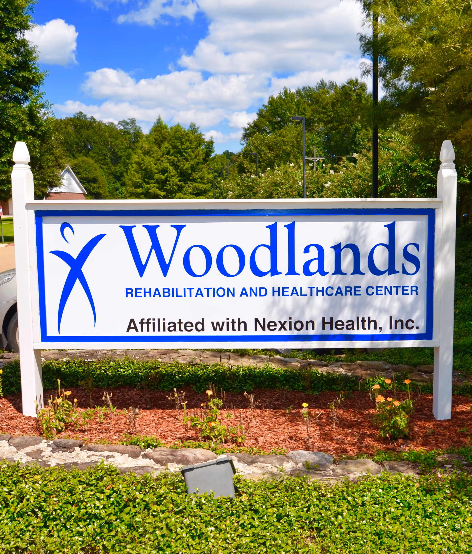 Outdoor sign for Woodlands Rehabilitation and Healthcare Center, affiliated with Nexion Health, Inc., surrounded by greenery and under a partly cloudy blue sky.