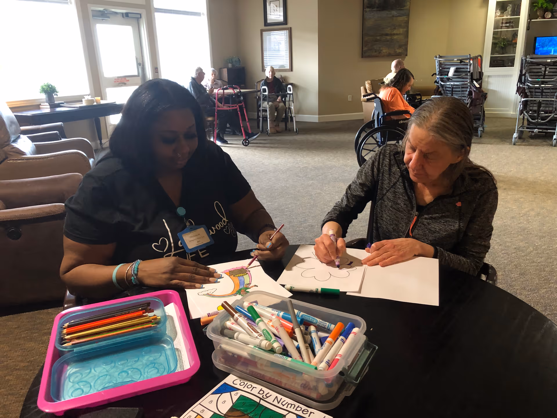 A caregiver and an elderly woman sitting at a round table engaged in coloring activities with markers and colored pencils in a common area of a senior living facility. Other elderly residents are seated in the background, some in wheelchairs and some using walkers.