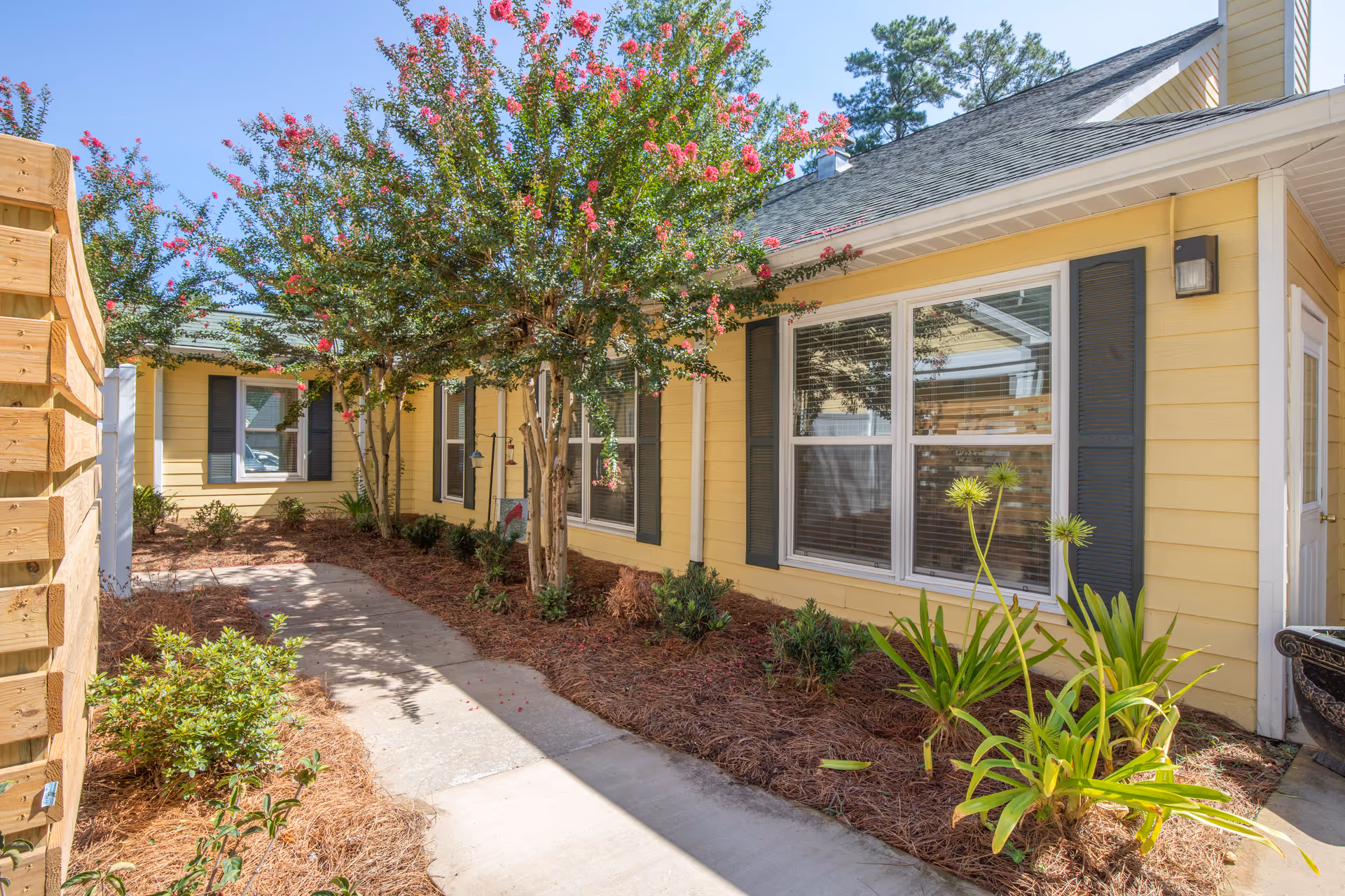 Walkway bordered by landscaping leading past a yellow single-story building with shuttered windows and flowering trees.