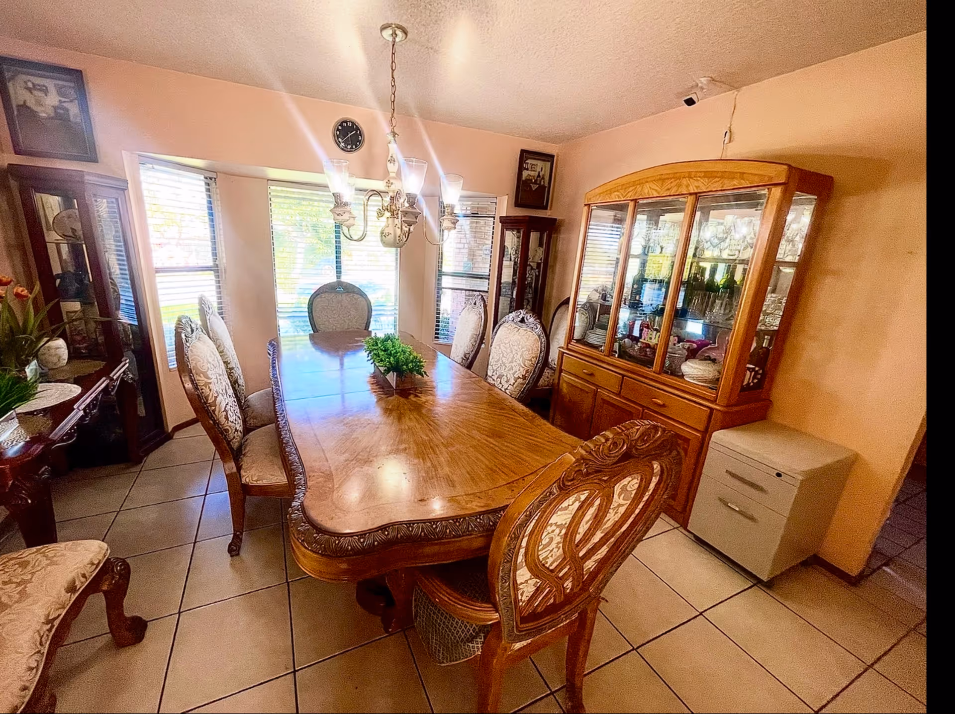 A traditional dining room with a large wooden dining table surrounded by ornate upholstered chairs. The room features a wooden china cabinet filled with glassware and dishes, a chandelier hanging above the table, and three windows with blinds letting in natural light. The walls are painted peach, and there is a small filing cabinet in the corner.