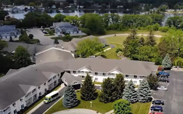 Aerial view of Charleston House Assisted Living facility showing a large two-story building with a gray roof surrounded by green trees and well-maintained lawns. There is a circular driveway with several parked cars and a small bus near the entrance. In the background, there are other buildings and a body of water.