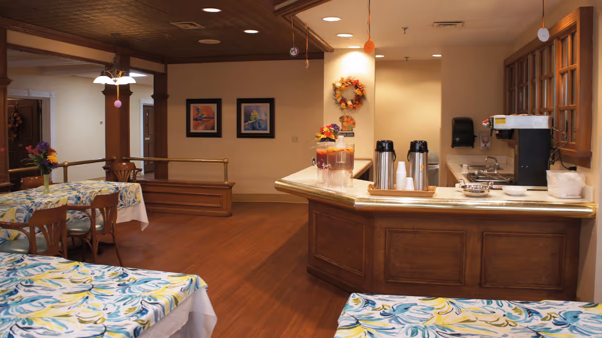 Interior view of a senior living facility dining area with tables covered in floral tablecloths and wooden chairs. A counter with coffee dispensers, cups, and a beverage container is visible on the right side. The room has wooden flooring, framed artwork on the walls, and a decorative wreath hanging on a column.