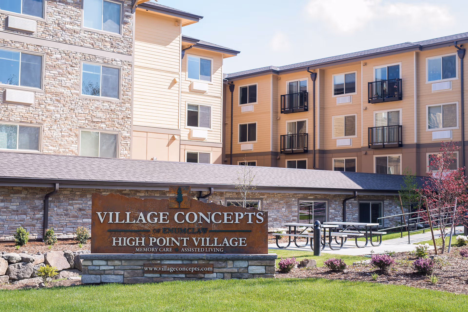 Exterior view of Village Concepts of Enumclaw - High Point Village, a senior living facility with a stone and beige siding facade, multiple windows, and small balconies. In front, there is a landscaped area with grass, shrubs, and a large sign displaying the facility name and website. Picnic tables are visible on a paved area near the building.
