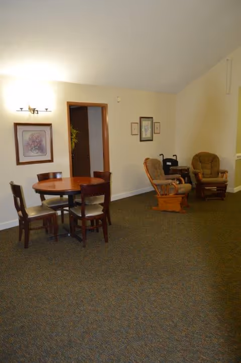 Communal room with a round wooden table and four chairs, two upholstered armchairs and a walker against a beige wall.