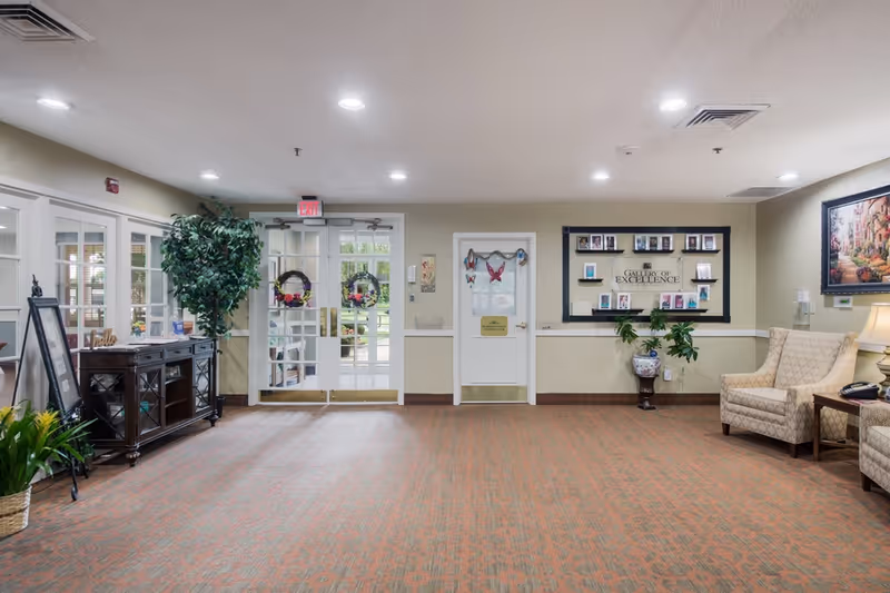 Bright senior living lobby with seating, decorative plants, a display shelving unit, and glass double doors leading outside.