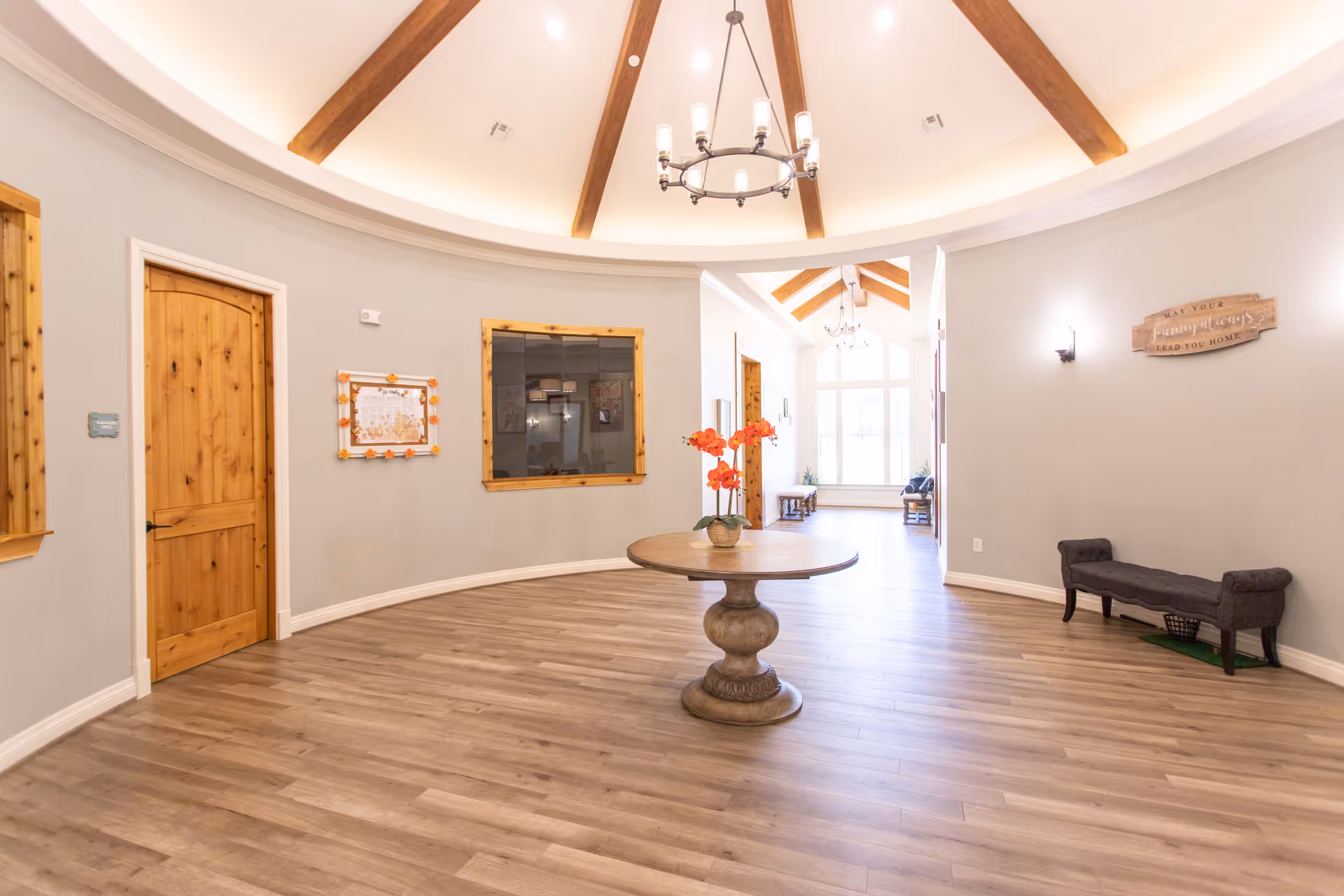 A spacious, well-lit interior room with a round wooden table in the center holding a vase with orange flowers. The room has light-colored walls, wooden doors and window frames, and a wooden floor. The ceiling is vaulted with exposed wooden beams and a chandelier hanging from the center. There is a bench along the right wall and a large window at the far end letting in natural light.