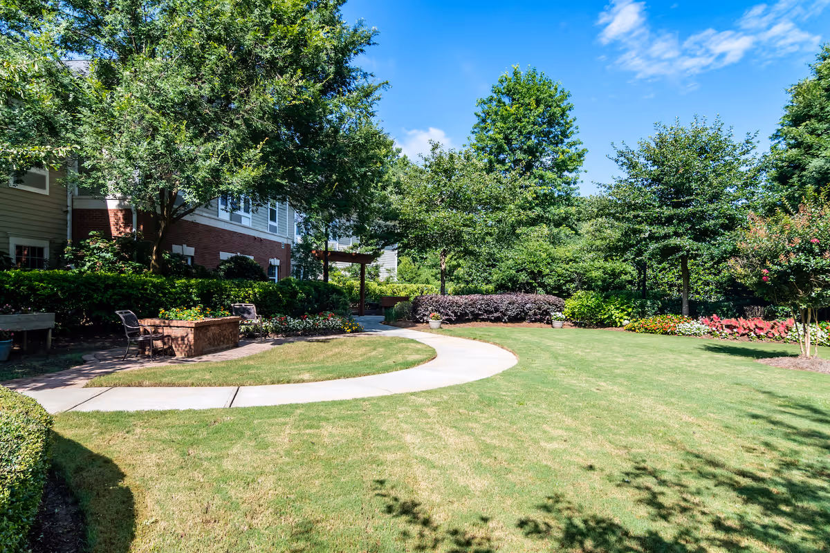 A well-maintained outdoor garden area with a curved concrete pathway, green grass, various trees, shrubs, and colorful flower beds. There are benches and a brick planter near the pathway, with a building partially visible on the left side under a bright blue sky.