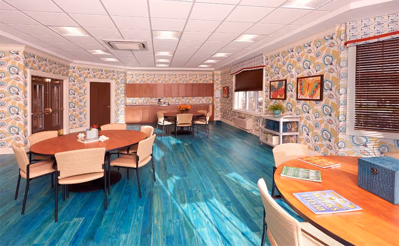 Bright communal dining/activity room with round wooden tables and chairs, floral wallpaper, and turquoise wood-look flooring.