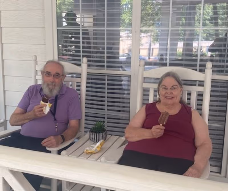 An elderly man and woman sitting on white rocking chairs on a porch, each holding a chocolate ice cream bar. There is a small table between them with a small potted plant and an empty ice cream wrapper. Behind them is a window with closed blinds.