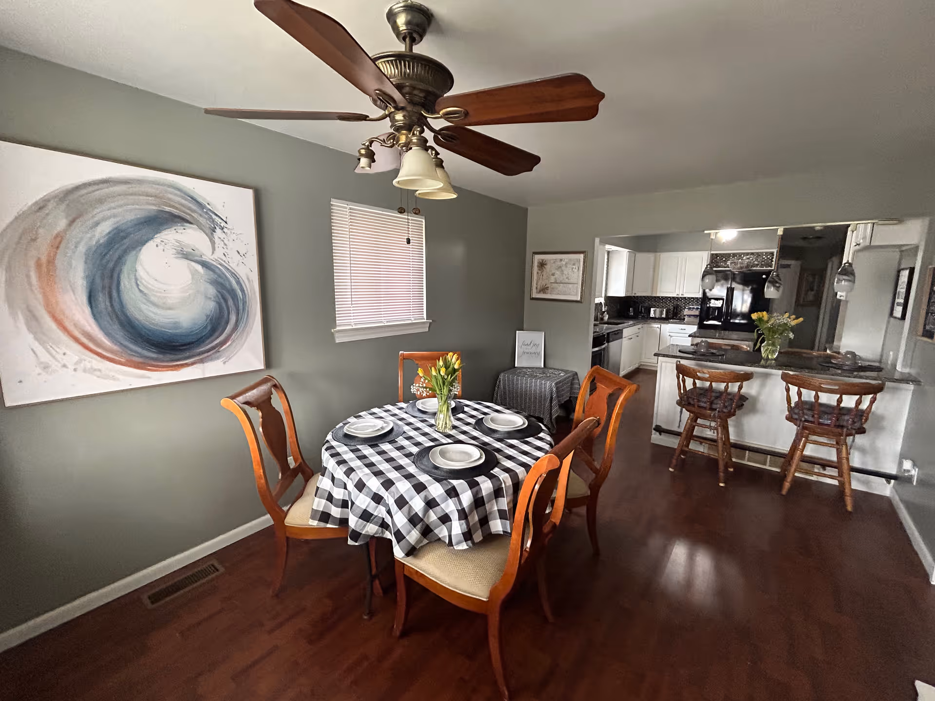 Dining area with a round table set with plates and a black-and-white checkered tablecloth, wooden chairs, a ceiling fan, and a view into a kitchen with bar stools.