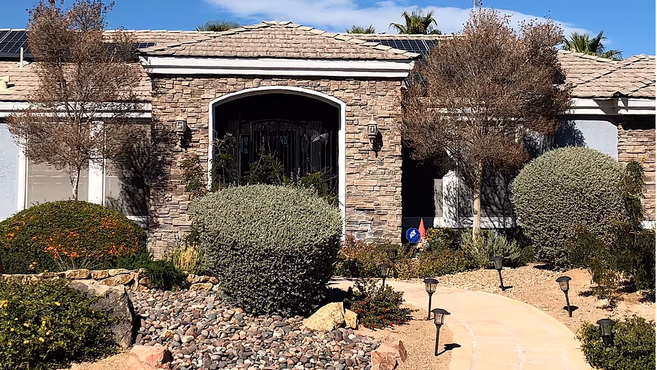 Front exterior view of a single-story building with stone facade and a tiled roof, surrounded by landscaped bushes, small trees, and a stone pathway with solar lights leading to the entrance under a clear blue sky.