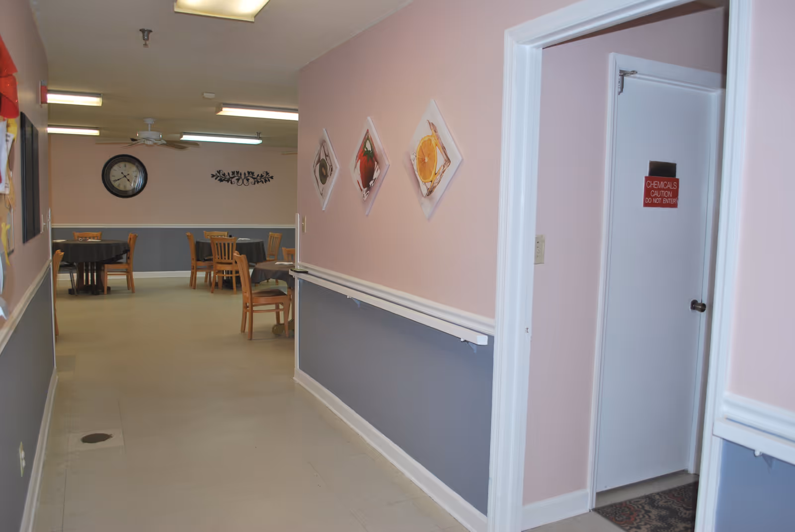 Hallway leading to a dining area with tables and chairs, pastel pink and gray walls, wall art, and a door labeled 'CHEMICALS CAUTION DO NOT ENTER'.