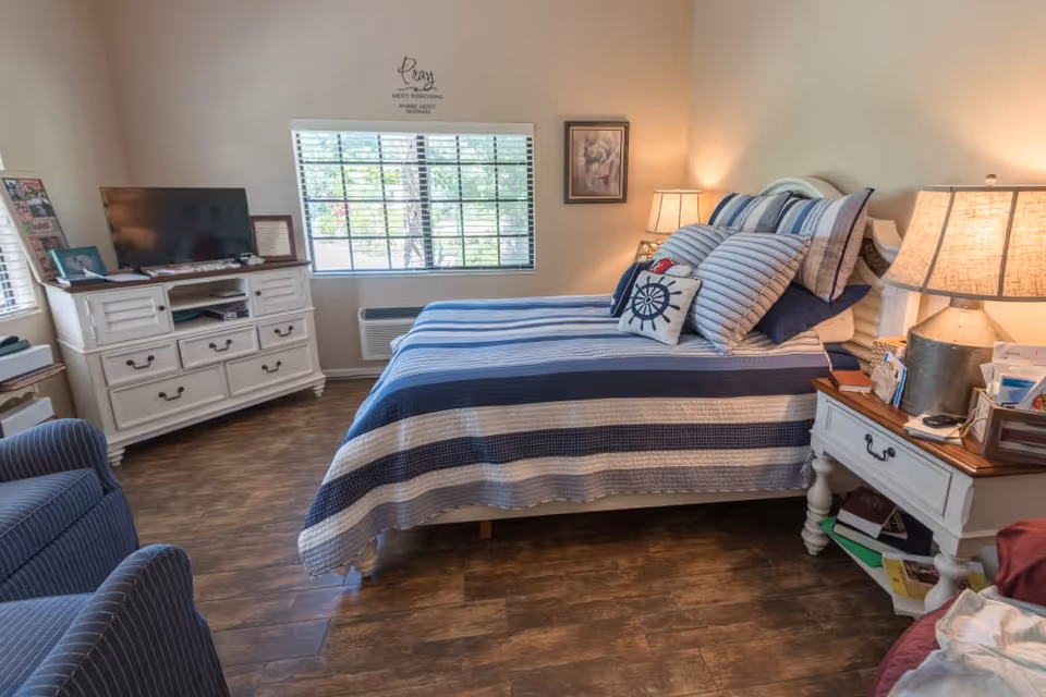 A cozy bedroom with a bed covered in a blue and white striped quilt and multiple pillows, including a decorative pillow with a nautical design. There is a white dresser with a TV on top, a window with blinds letting in natural light, a framed picture on the wall, and two bedside tables with lamps. The floor is wooden, and there is a blue armchair partially visible in the foreground.