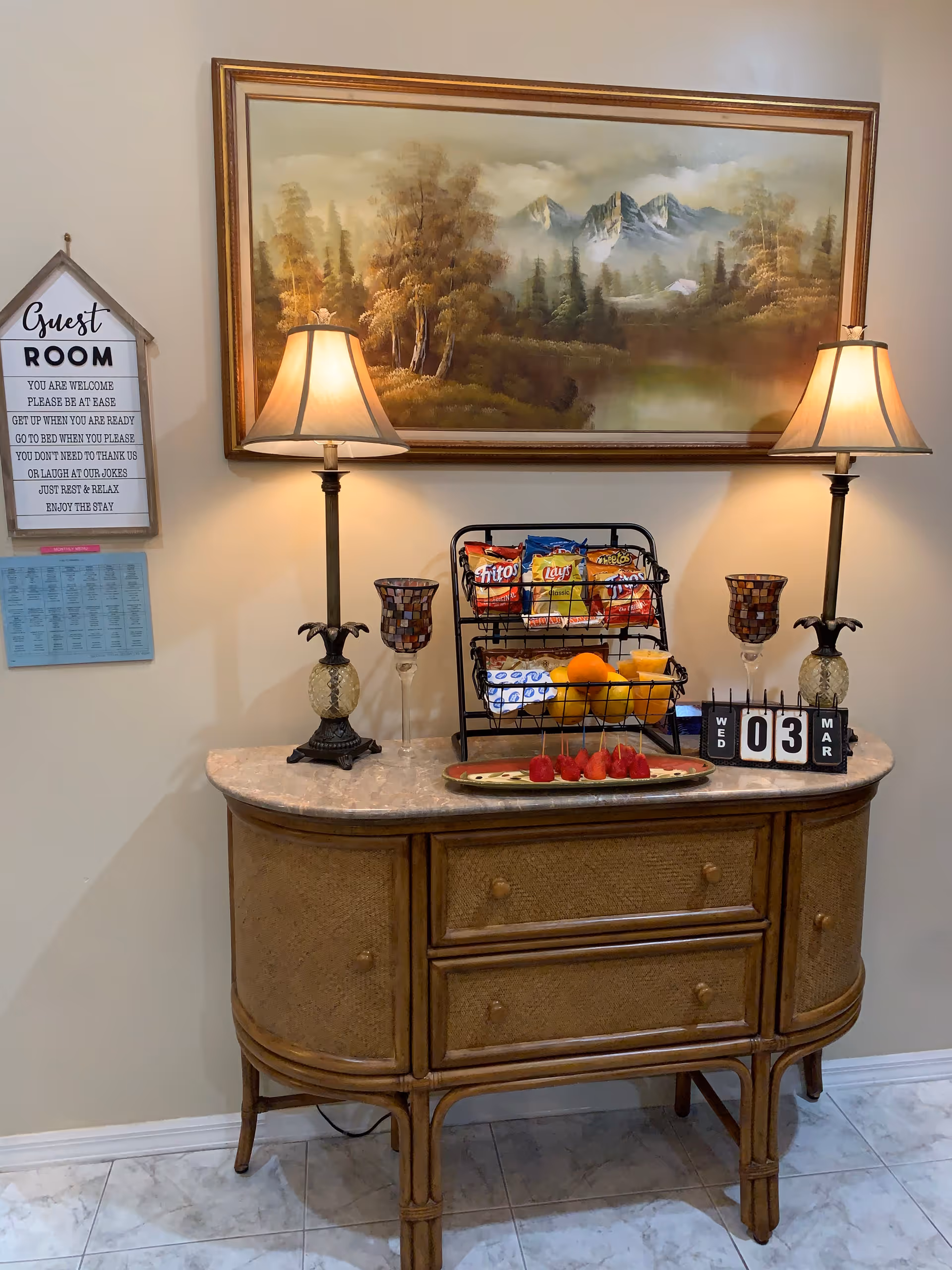 Decorative console table topped with two lamps, a snack rack, fruit, and decor beneath a large landscape painting in an interior entry area.