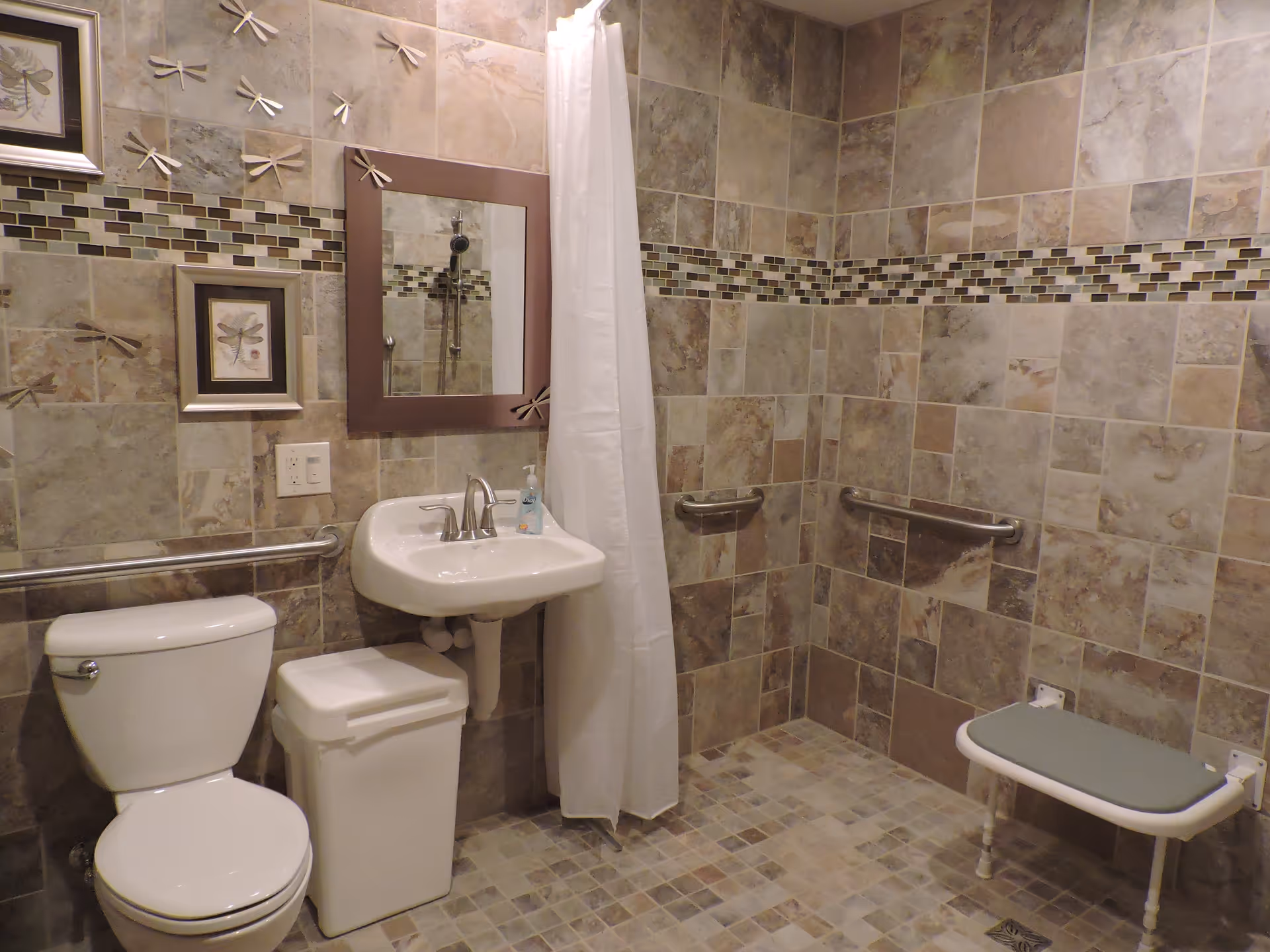 Accessible tiled bathroom with a toilet, wall-mounted sink, shower curtain, grab bars and a fold-down shower seat.