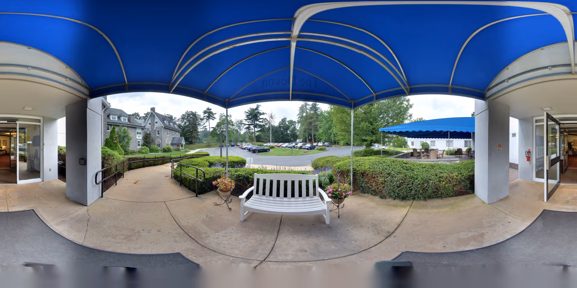 Outdoor covered entrance area at The Devon Senior Living with a white bench and potted flowers under a blue canopy. There are two open doorways leading inside the building on either side, landscaped bushes, a parking lot with cars, and trees in the background.