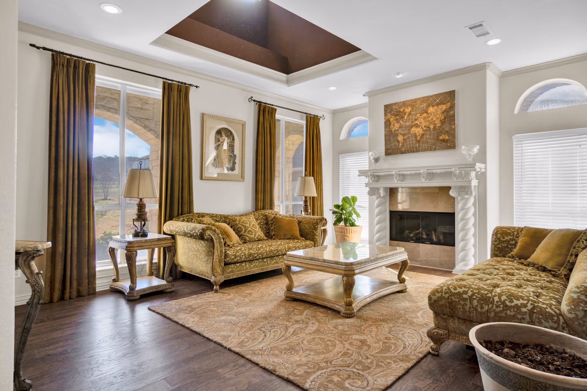 A well-lit living room with two ornate gold-patterned sofas, a matching coffee table, and side tables with lamps. The room features large windows with brown curtains, a decorative fireplace with a world map artwork above it, and a beige patterned rug on dark wooden floors.