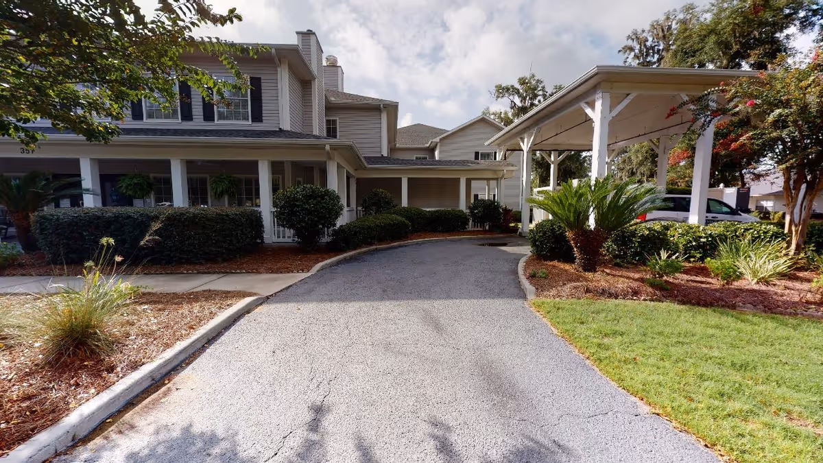 Exterior view of a senior living facility named Summer Breeze Senior Living, showing a paved driveway leading to a covered entrance with white pillars. The building is two stories with beige siding and black shutters. There are well-maintained bushes, plants, and a green lawn surrounding the driveway under a partly cloudy sky.