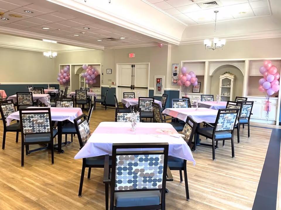 A dining room with multiple tables covered in light purple tablecloths and decorated with small centerpieces. The chairs have patterned backs with circles in shades of blue, gray, and beige. The room has wooden flooring, light-colored walls with blue wainscoting, and ceiling lights. Pink and purple balloons are arranged in clusters near the walls.