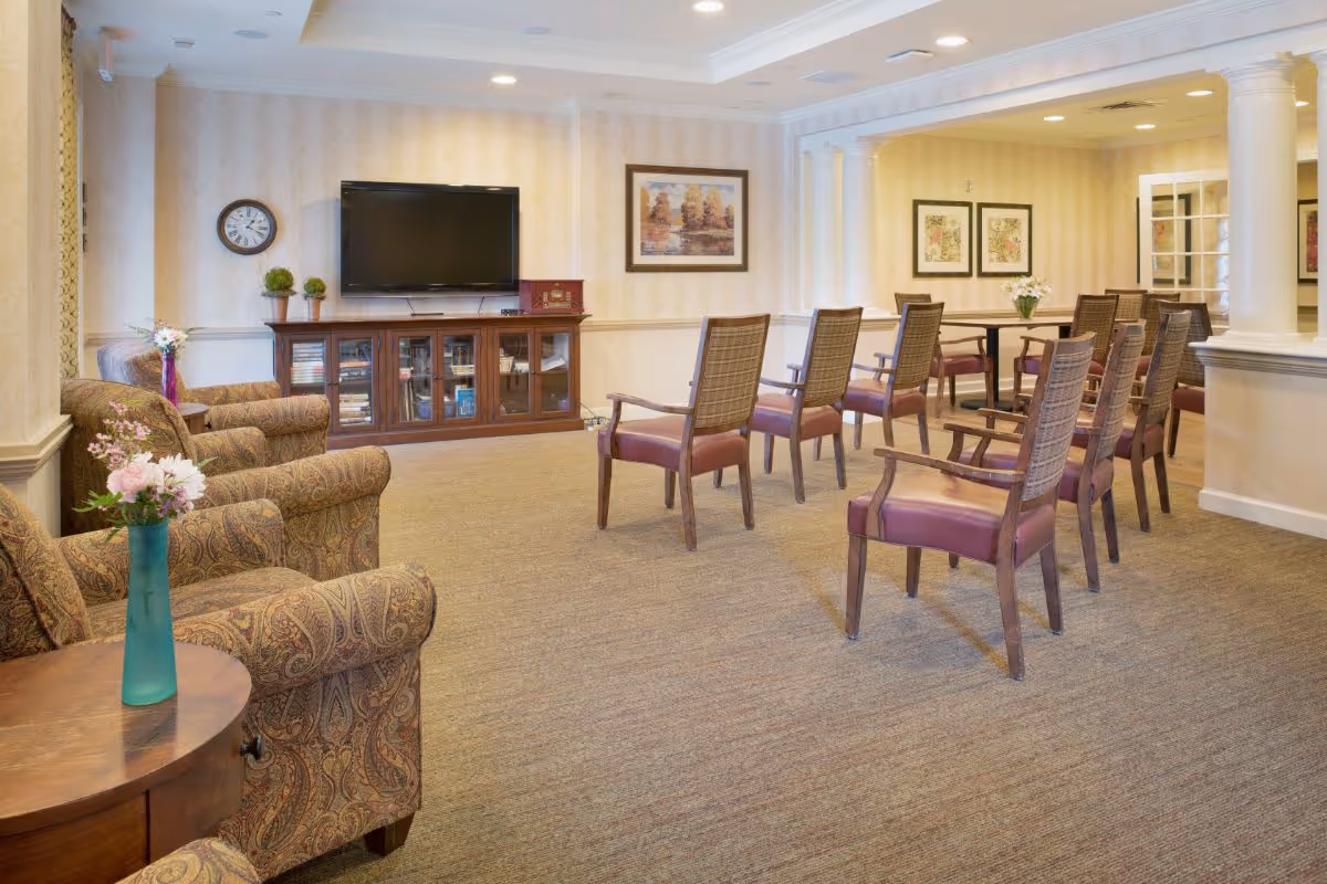 A bright communal lounge with upholstered armchairs and rows of wooden chairs facing a wall-mounted TV and media cabinet.