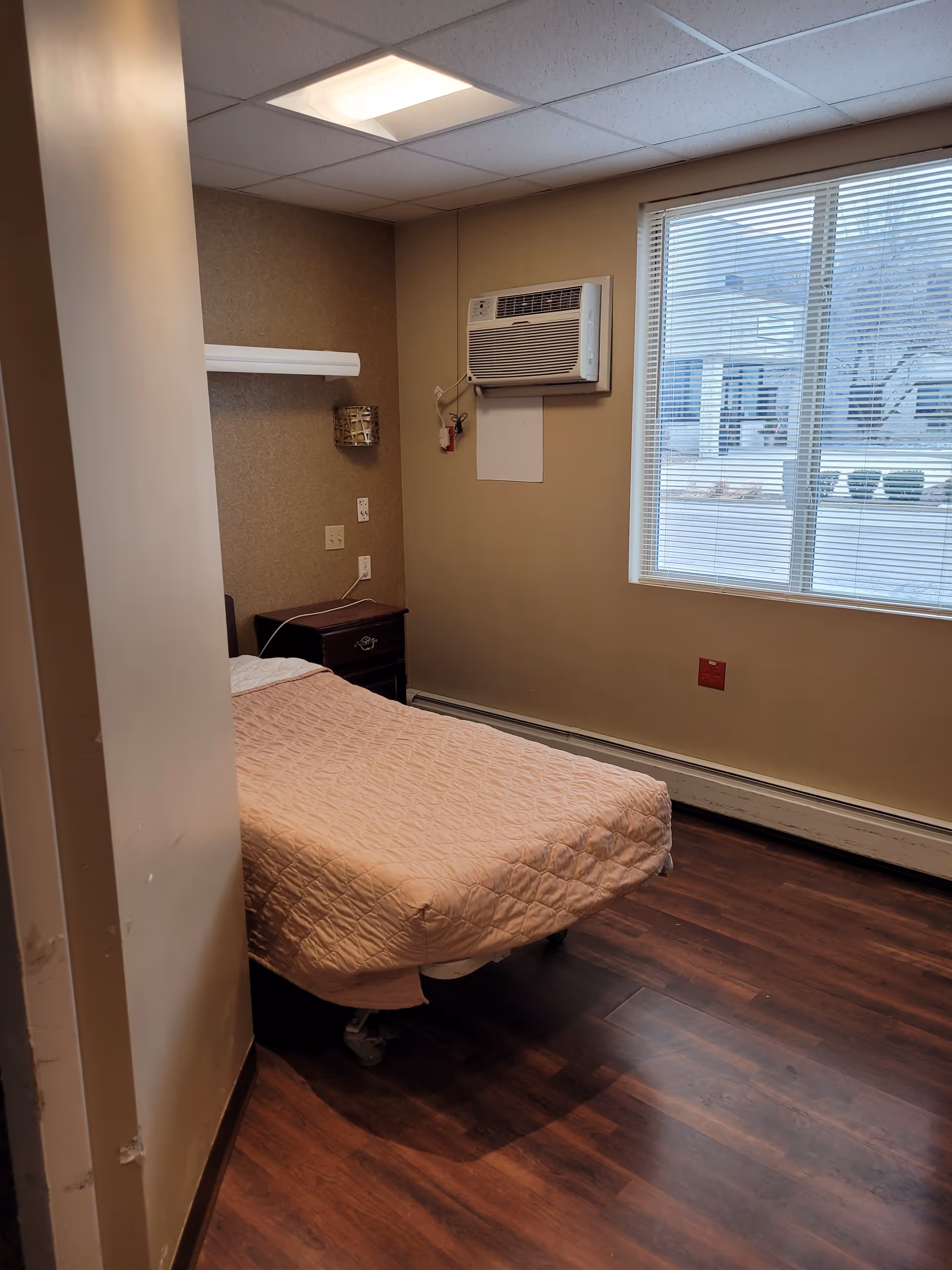 A small bedroom with a single bed covered in a light pink quilt. There is a wooden nightstand next to the bed, a wall-mounted air conditioning unit above it, and a window with blinds letting in natural light. The floor is dark wood, and the walls are beige.