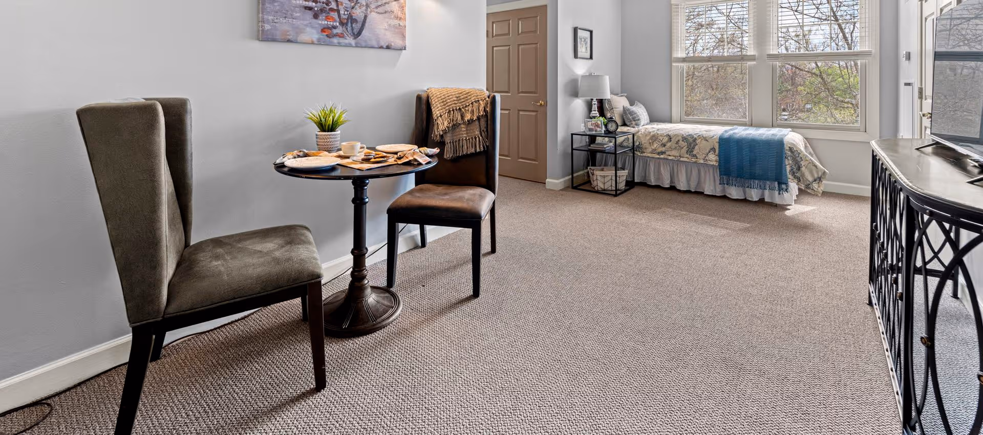 A cozy room with a small round table set with plates and cups, two upholstered chairs, a single bed with patterned bedding and a blue throw, a bedside table with a lamp and framed photos, large windows letting in natural light, and a TV on a decorative stand.