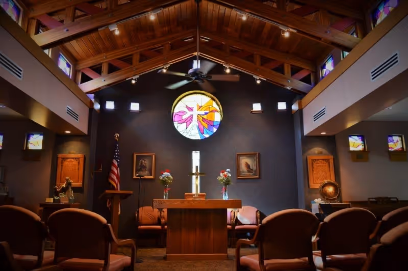 Interior of a chapel with wooden vaulted ceiling, stained glass windows including a large circular one with a cross design, an altar with a cross and flower vases, chairs arranged facing the altar, and framed pictures on the walls.
