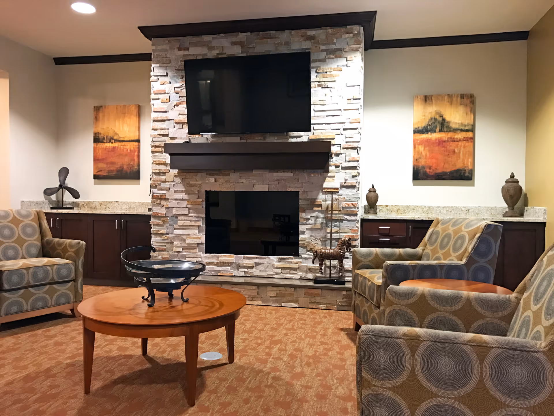 A cozy living room area with three patterned armchairs arranged around a wooden coffee table. The room features a stone fireplace with a mounted flat-screen TV above it. On either side of the fireplace are dark wood cabinets topped with granite countertops, decorated with vases and abstract paintings on the walls.
