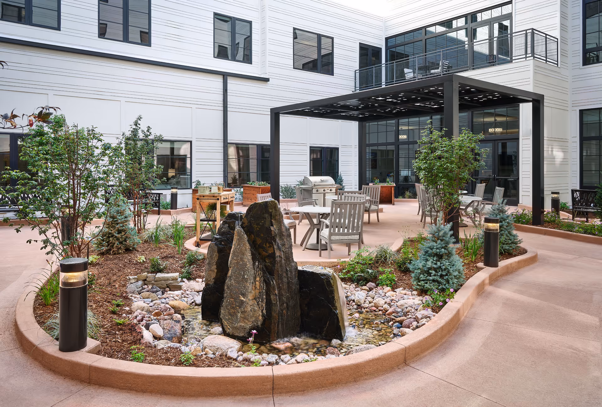 Outdoor courtyard area at MorningStar Senior Living at Old Town featuring a small rock water fountain surrounded by plants and shrubs. There are several seating areas with tables and chairs under a pergola, and the courtyard is enclosed by the building with multiple windows overlooking the space.