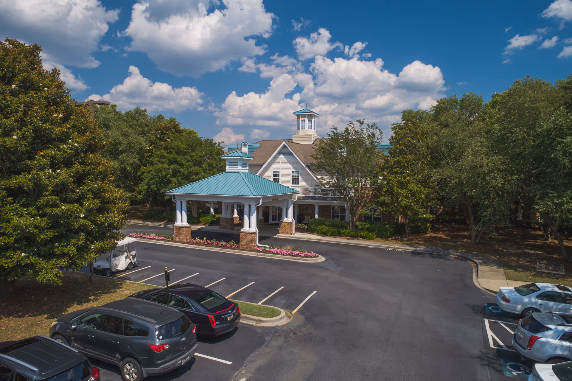 Exterior view of Wildewood Downs facility showing a building with a covered entrance, surrounded by trees and a parking lot with several cars parked. The sky is partly cloudy with blue patches.