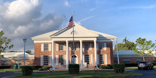 Front exterior view of a large brick building with white columns and an American flag on a flagpole in front. The building has multiple windows and a triangular pediment above the entrance. There are bushes and a lawn in front of the building, with a clear blue sky and some clouds in the background.