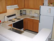 Small kitchen area with wooden cabinets, a white refrigerator, an electric stove with four burners, a white range hood, a sink with a faucet, and various kitchen items on the countertops including a coffee maker and a framed picture.