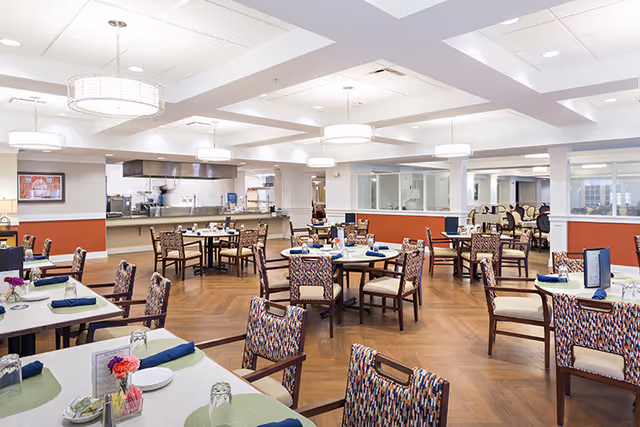 A spacious dining room with multiple tables and chairs arranged neatly. Each table is set with plates, glasses, napkins, and small flower arrangements. The room features wooden flooring, modern ceiling lights, and an open kitchen area in the background.