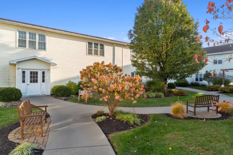 Outdoor garden area at The Hearth at Greenpoint featuring a paved walkway, two wooden benches on small brick patios, green grass, a flowering shrub with pink blooms, and a large tree. The background shows a white two-story building with multiple windows and a white door under a small awning.