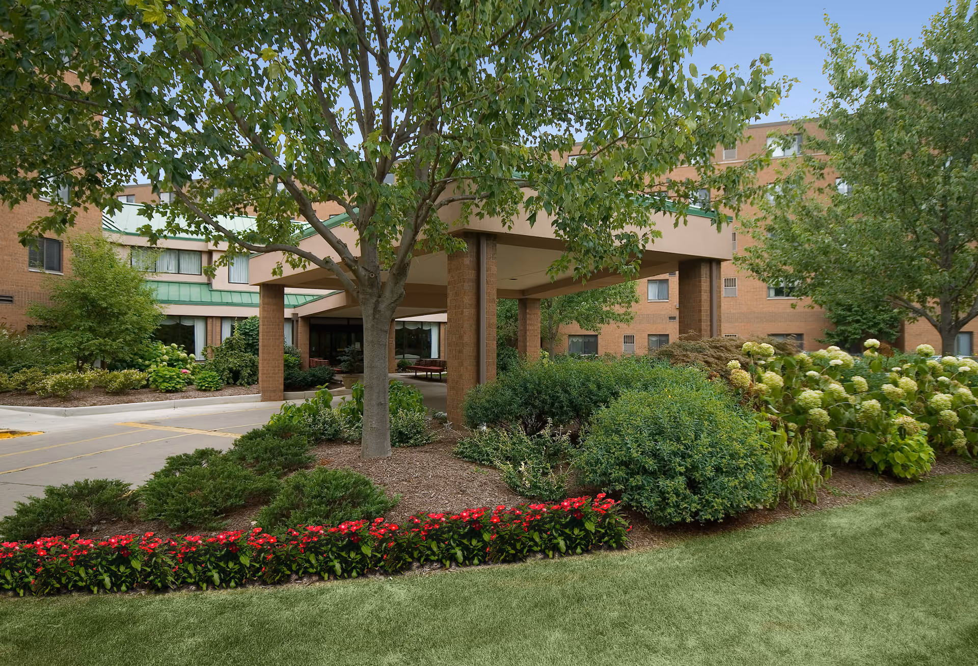 Entrance area of Ohio Living Swan Creek facility showing a covered drop-off with brick pillars surrounded by well-maintained landscaping including green bushes, trees, and a row of red flowers in front of a multi-story brick building under a clear blue sky.