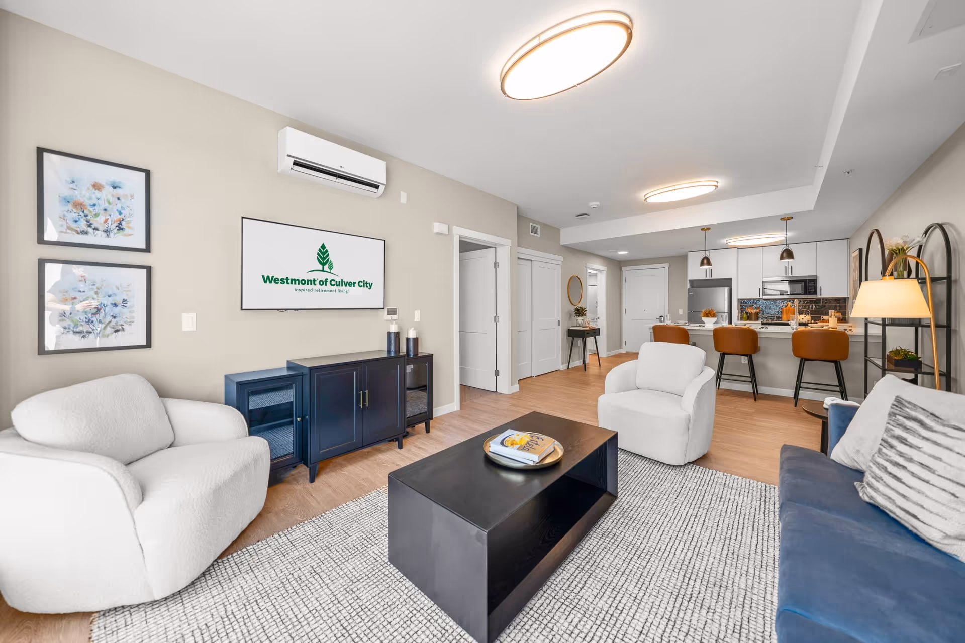 A modern, open-concept living room and kitchen area in a senior living facility. The living room features a white armchair, a blue sofa with a striped pillow, a black coffee table on a patterned rug, and a black cabinet under a wall-mounted TV displaying the Westmont of Culver City logo. The kitchen has a breakfast bar with three brown bar stools, white cabinetry, stainless steel appliances, and pendant lighting. The walls are light beige, and there are two framed floral artworks on the wall near the TV.