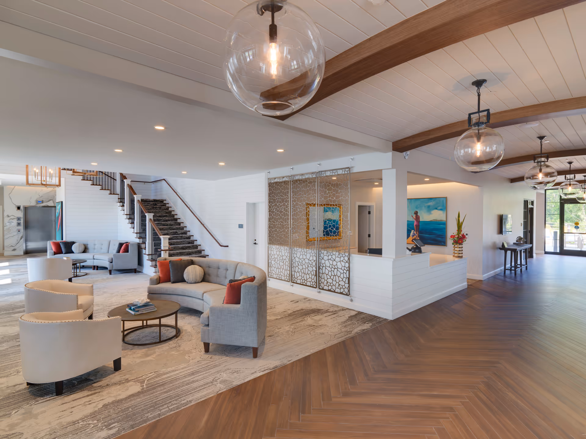 A spacious and modern senior living facility lobby with a seating area featuring curved and straight gray sofas, beige armchairs, and round coffee tables. The room has wooden beams on the ceiling, large glass pendant lights, a staircase with a carpet runner, and decorative wall panels. There is a reception desk with a vase of flowers and a colorful painting on the wall behind it. The floor is a combination of wood and carpet, and large windows allow natural light to fill the space.