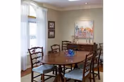 Dining room with a wooden oval table surrounded by six chairs, a sideboard, artwork on the wall, and a window with sheer curtains.