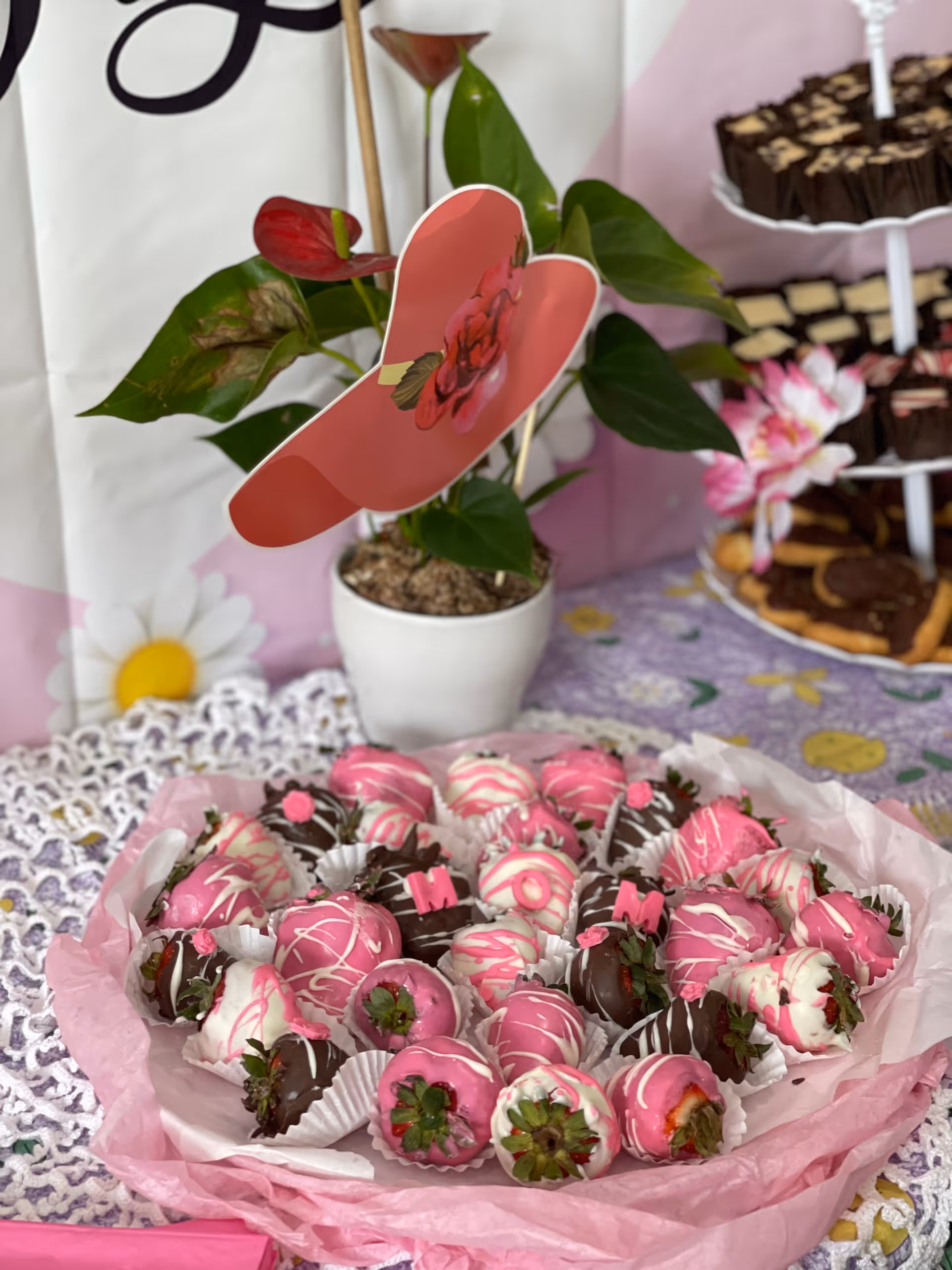 A decorative plate filled with strawberries dipped in pink, white, and dark chocolate, some decorated with pink icing letters. Behind the plate is a white potted plant with green leaves and a red paper butterfly decoration. In the background, there is a tiered tray with various chocolate treats and a floral tablecloth with a lace doily.